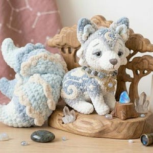 May include: A handcrafted crochet wolf plush with blue, white, and cream yarn, featuring a star-patterned design, a beaded collar with a star charm, and a large fluffy tail. It sits on a wooden base with a card reading DREAMWEAVER CLOUD WOLF - CROCHET PATTERN.
