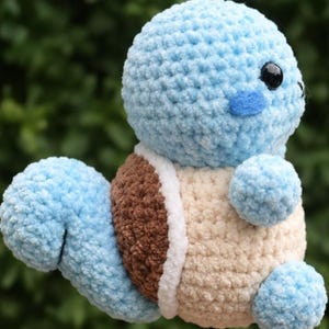 Può includere: Un peluche fatto a mano all'uncinetto a forma di personaggio dei cartoni animati. Il giocattolo è principalmente azzurro, con un guscio marrone, pancia color crema e occhi neri. Il peluche ha una testa rotonda, braccia piccole e una coda.