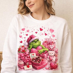 May include: White sweatshirt featuring a Grinch character in a Santa hat surrounded by Valentine's Day gifts and hearts. The Grinch holds a heart-shaped sign that says "I ❤️ YOU". A teddy bear and gifts are also visible.