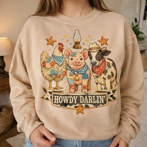 Peut inclure: Sweat-shirt beige avec un dessin d'un poulet, d'un cochon et d'une vache de dessin animé, portant des bandanas et des chapeaux. Le cochon a une cloche. Le texte "HOWDY DARLIN'" est sur une bannière. Des étoiles et une lanterne sont également présentes.