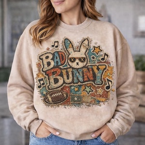 May include: Beige sweatshirt featuring a graphic of a bunny wearing sunglasses with the text "BAD BUNNY" in colorful letters. The design includes musical notes, stars, and a football. The sweatshirt has long sleeves and a crew neck.
