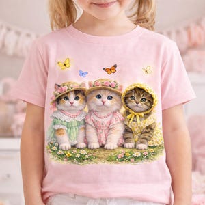 May include: Pink t-shirt featuring three adorable kittens wearing bonnets and dresses. The kittens are surrounded by butterflies and flowers. The kittens are wearing a green, pink, and yellow dress. The t-shirt is made of a soft material.