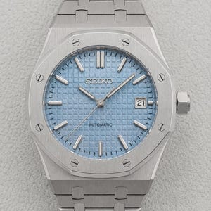 May include: A silver-toned stainless steel wristwatch featuring an octagonal bezel with eight visible screws. The watch has a light blue textured waffle-pattern dial, silver baton indices, silver hands, and a date window displaying the number 21.
