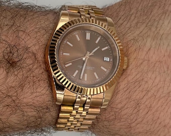 May include: A gold-toned wristwatch with a brown dial and a date display at the 3 o'clock position. The watch features a fluted bezel and a gold link bracelet. The watch is shown on a wrist.