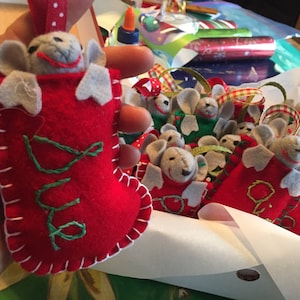 May include: A collection of felt mouse ornaments, each with a different name embroidered on the stocking. The mice are peeking out of the stockings, which are red, green, and white.