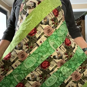 May include: A green and red patterned apron with a diagonal design. The apron has a green fabric with a floral pattern and a green fabric with a paisley pattern. The apron is made of cotton fabric.