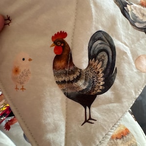 May include: A close-up of a quilted fabric featuring a rooster and chick design. The rooster has brown, black, and red feathers. The chick is yellow. The background is a light beige color with other chicken illustrations.