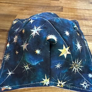 May include: A navy blue bowl cozy with a celestial design featuring stars and a crescent moon. The fabric is patterned with white, yellow, and gold stars against a dark blue background. The cozy is quilted and has a white stitched border.
