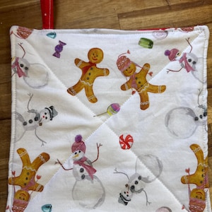 May include: White quilted pot holder with a red loop. The fabric features a repeating pattern of gingerbread people, snowmen, and candies in various colors. The pot holder is square with a stitched design.