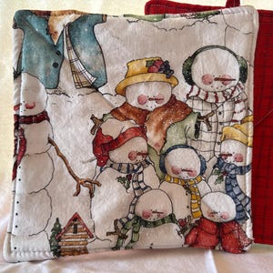 May include: Set of two white cotton pot holders with a winter snowman theme. The pot holders feature a variety of snowmen with different hats, scarves, and accessories. The background is a white snowy scene with a small wooden house.