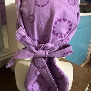 May include: Purple fabric bandana with a pattern of circles of small hearts. The bandana is tied around a mannequin's head.