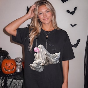 May include: A black short-sleeve t-shirt featuring a graphic print of a white ghost sitting on a swing. The ghost is blowing a pink bubble with bubblegum. The design includes two small black bats flying near the ghost and silver chains.