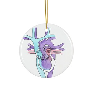 Puede incluir: Adorno redondo de cerámica blanca con una ilustración en acuarela de un corazón humano y las venas circundantes. El corazón es morado, con venas azules y rosas. Se adjunta un hilo dorado para colgarlo.