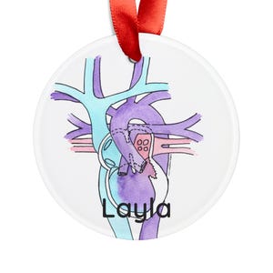 Puede incluir: Adorno redondo blanco con una ilustración de acuarela de un corazón y venas en tonos morado, azul y rosa. El nombre "Layla" está escrito en negro en la parte inferior. Se adjunta una cinta roja para colgar.