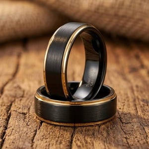Puede incluir: Dos anillos negros de carburo de tungsteno con una banda interior y un borde exterior de color dorado. Los anillos tienen un acabado de metal cepillado y se muestran sobre una superficie de madera. Un anillo está apilado sobre el otro.