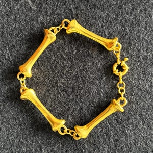 May include: Gold-toned bracelet featuring a bone design. The bracelet is composed of multiple bone-shaped links connected by small chain segments. The clasp is a circular design. The jewelry is displayed on a dark gray surface.