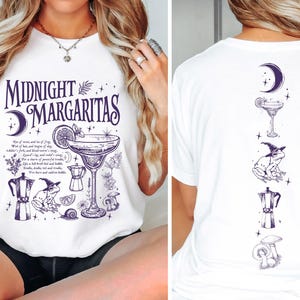 May include: A white t-shirt featuring a dark purple graphic print. The front displays the text "MIDNIGHT MARGARITAS" with illustrations of a margarita glass, a frog in a witch hat, a moka pot, mushrooms, and a crescent moon. The back shows a vertical alignment of these same icons.