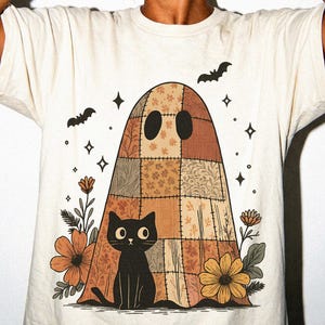 Puede incluir: Una camiseta de color crema con un gráfico de un fantasma hecho de tela de retazos marrón y bronceada. Un gato negro se sienta frente al fantasma, rodeado de flores naranjas y amarillas, estrellas pequeñas y dos murciélagos negros de fondo.