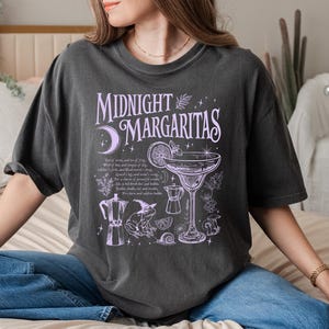 May include: Dark gray t-shirt with the text "Midnight Margaritas" in lavender. The shirt features a margarita glass, a coffee pot, and other illustrations. The shirt is a comfortable, oversized fit.