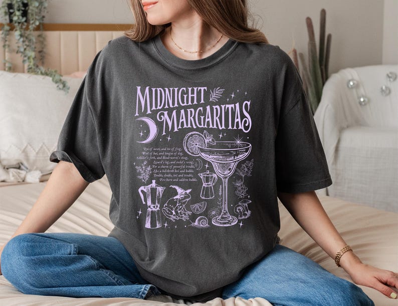May include: Dark gray t-shirt with the text "Midnight Margaritas" in lavender. The shirt features a margarita glass, a coffee pot, and other illustrations. The shirt is a comfortable, oversized fit.