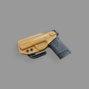 May include: A tan leather holster with a black handgun inside. The holster has a clip on the side for attaching to a belt.