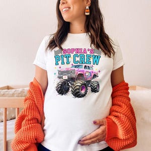 May include: White t-shirt featuring a pink monster truck graphic with the text "SOPHIA'S PIT CREW" in blue and pink. The shirt has a crew neck and short sleeves. The design includes a checkered flag and a unicorn.