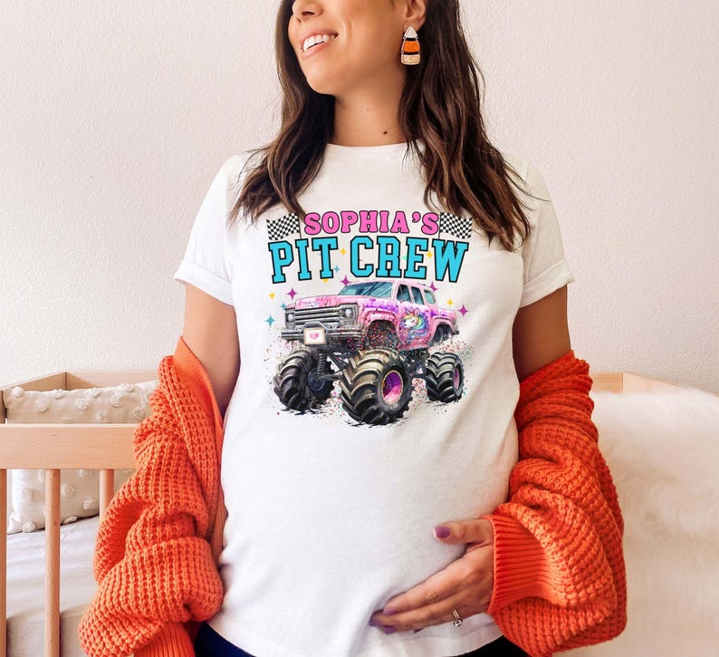May include: White t-shirt featuring a pink monster truck graphic with the text "SOPHIA'S PIT CREW" in blue and pink. The shirt has a crew neck and short sleeves. The design includes a checkered flag and a unicorn.