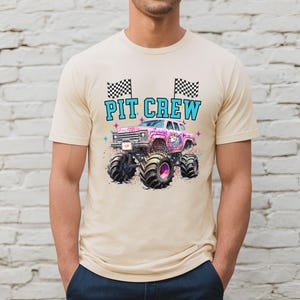 May include: Beige t-shirt featuring a colorful graphic of a monster truck with the text "PIT CREW". The monster truck is pink and purple with large black tires. Two checkered flags are above the text.