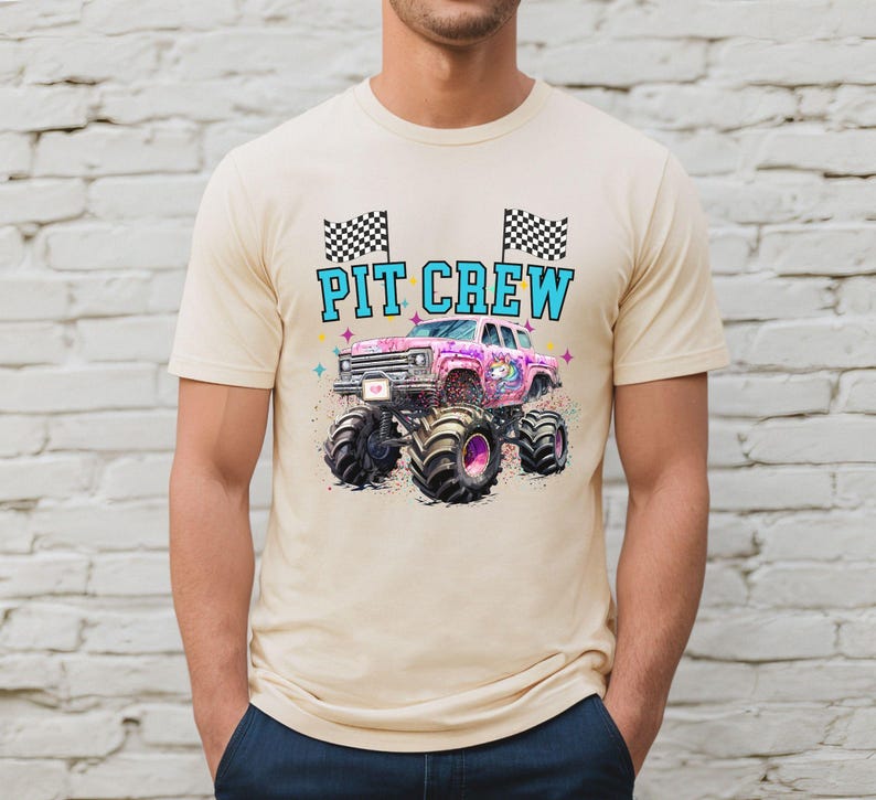 May include: Beige t-shirt featuring a colorful graphic of a monster truck with the text "PIT CREW". The monster truck is pink and purple with large black tires. Two checkered flags are above the text.
