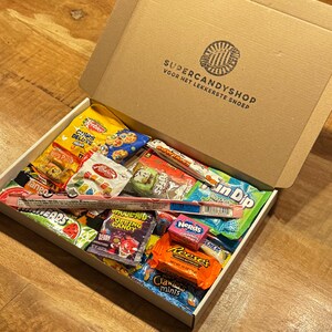 Può includere: Una scatola di cartone piena di una varietà di caramelle colorate, tra cui Nerds, Reese's Pieces e Sour Patch Kids. La scatola è etichettata "Supercandyshop" e "Voor het lekkerste snoep".