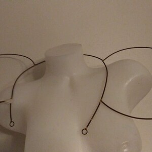 以下が含まれることがあります： 白いマネキンに展示された、3つの円形要素を持つワイヤーフレームのヘッドピース。濃い色の金属の円は細いワイヤーで接続されており、ミニマリストで抽象的なデザインになっています。ヘッドピースは頭と肩に着用するように設計されています。