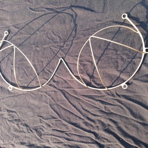 May include: A wireframe bra in a minimalist style. The bra is constructed from thin, white metal wires, forming the cups and underbust band. The design features a simple, geometric aesthetic, with circular and triangular shapes. The bra is laid flat on a dark, textured surface.