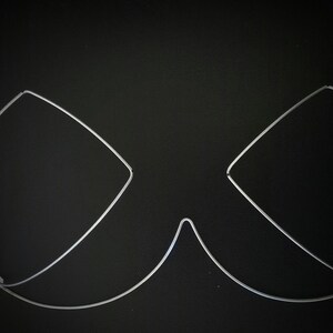 May include: A silver-colored wireframe bra, composed of curved and angular lines, rests against a black background. The design features two cup shapes and a connecting underbust wire. Small loops are present at the ends of the wires.