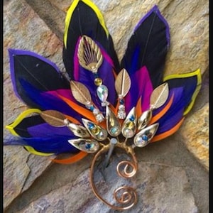 Könnte beinhalten: Ein handgefertigter Ohrschmuck mit Federn in Schwarz, Lila, Orange und Gelb. Das Design umfasst goldfarbene Blätter und glitzernde Edelsteine, mit einem Ohrbügel aus Kupferdraht.