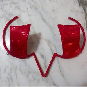 Puede incluir: Un top de sujetador rojo con lentejuelas, con una parte inferior en forma de V y correas curvas. Las copas del sujetador son rectangulares y están conectadas por un fino cordón rojo. Las correas tienen pequeños bucles en los extremos.