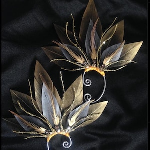 May include: Two ear cuffs with a leaf and feather design. The leaves are gold and brown, with grey and brown feathers. The cuffs have silver wire accents and small beads. The background is black.
