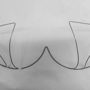 May include: A grayscale image of a wireframe bra, constructed from thin, silver-colored metal. The bra's design includes two cups and a central bridge, with curved underwire and straps. The structure is set against a plain white background.