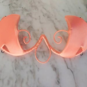 May include: A pair of peach-colored eye mask with a whimsical design featuring two curled wire accents.