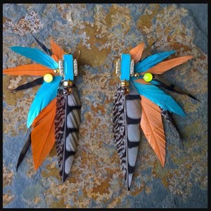 Op de afbeelding: Een paar handgemaakte oorringen met veren. Elke oorring heeft een rechthoekige turkooizen steen, goudkleurige kralen en een cluster van kleurrijke veren in blauw, oranje, zwart en grijs. De oorbellen zijn ontworpen in een bohemien stijl.