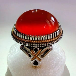 May include: A large, oval-shaped ring with a vibrant red gemstone. The ring features a silver band with gold accents and intricate detailing around the stone. The gemstone is polished and smooth, reflecting light.