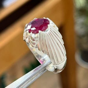 以下が含まれることがあります： 大きな楕円形のルビーの宝石が付いたシルバーリング。リングのセッティングは、ルビーの周りに詳細な扇形のデザインが施されています。リングは銀色のピンセットで保持されており、ジュエリーの複雑な職人技が際立っています。