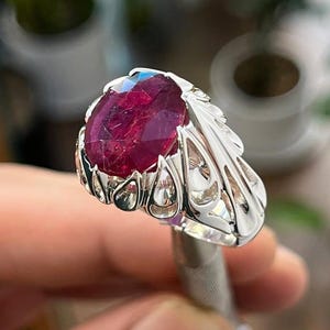 Peut inclure: Une bague en argent ornée d'une grosse pierre précieuse rouge foncé de forme ovale. Le sertissage de la bague présente un design ajouré orné. La pierre précieuse est le point central, captant la lumière et mettant en valeur sa couleur riche.