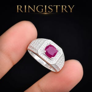 Peut inclure: Une bague en argent avec une pierre précieuse centrale rose foncé taille coussin entourée d'un halo de petites pierres blanches. L'anneau est large et incrusté de plusieurs rangées de minuscules pierres blanches. Le texte RINGISTRY est visible.