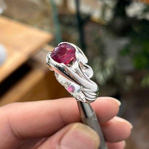 Op de afbeelding: Een zilveren ring met een grote, ovaal geslepen robijn. De zetting van de ring heeft een vloeiend, organisch ontwerp. De robijn heeft een diepe, levendige rode kleur en het zilver is hoogglans gepolijst.
