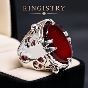 Puede incluir: Un anillo plateado con una piedra grande, ovalada y de color rojo intenso. El anillo tiene un diseño calado y ornamentado en los laterales y el texto "RINGISTRY" en la parte superior. El anillo se muestra en una caja de joyería negra.