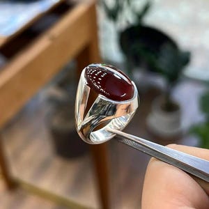 Könnte beinhalten: Ein polierter Silberring mit einem glatten, ovalen, tiefroten Edelstein in einer hohen Fassung. Die Ringschiene hat ein geteiltes Design mit offenen Seiten, das eine reflektierende metallische Oberfläche und einen dunklen, glänzenden Stein zeigt.