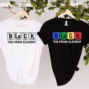 May include: Two t-shirts, one white and one black, hanging on wooden hangers. Both shirts feature the word "BLACK" in a periodic table style, with each letter in a different color. Below the word is the text "THE PRIME ELEMENT."