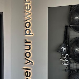 May include: A wall with the words "feel your power" in black, illuminated letters. The wall is white with a gray section on the right, where exercise equipment is stored.