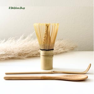 May include: A bamboo matcha tea whisk, scoop, and spoon set. The whisk has many thin tines and a black band. The scoop is long and curved, and the spoon is flat. The set is made of light brown bamboo.