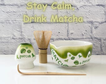 May include: A matcha tea set with a white ceramic bowl and a green rim, a bamboo whisk, a matcha bowl, and a bamboo scoop. The text "Stay Calm, Drink Matcha" is above the set.